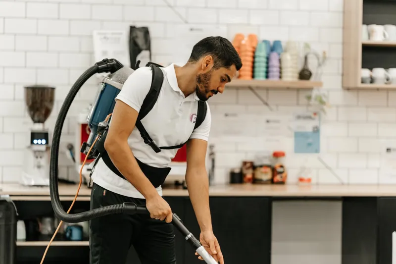 F&L cleaner in a branded white polo vacuuming an office kitchen with a backpack vacuum
